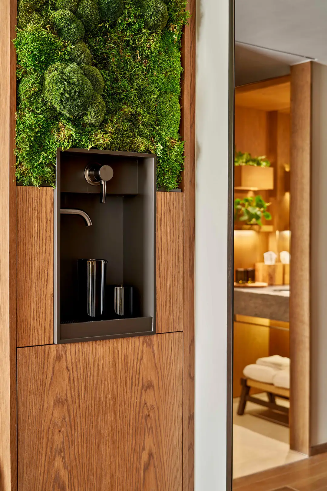 In-room water tap set into an oak wall niche below a moss panel, with a dark carafe and cup on the drip tray