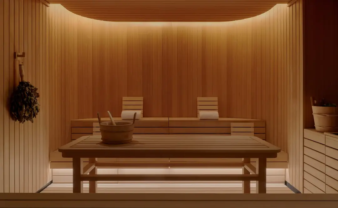 Wood-clad banya sauna with slatted benches, a wooden bucket on a table and a birch venik hanging on the wall