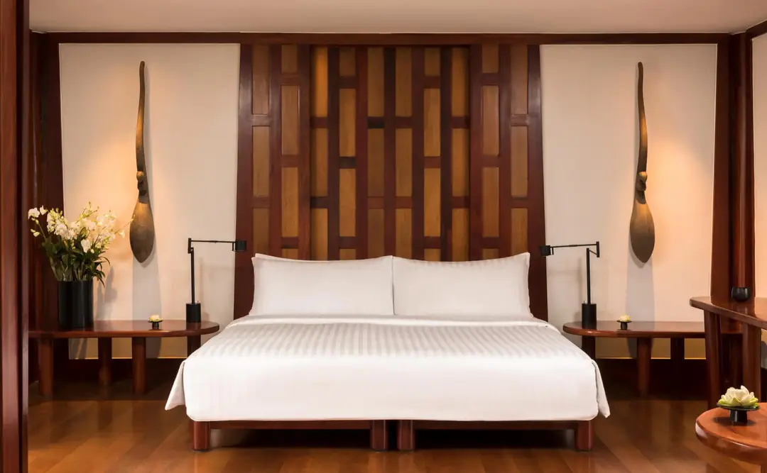 Pavilion bedroom with a white bed against a slatted teak headboard, wooden floor and bedside lamps