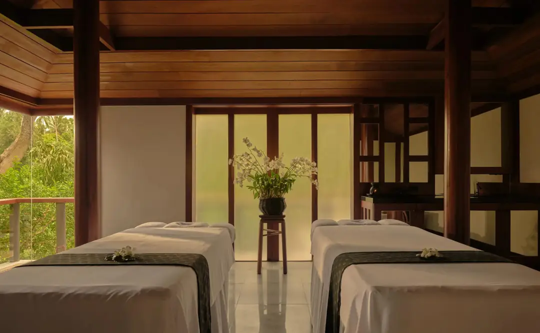Spa treatment room with two massage tables under a timber ceiling, orchids and a window onto trees