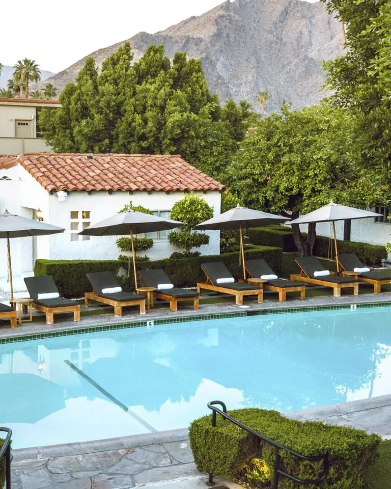 Pool with dark loungers and umbrellas beside a white tile-roofed bungalow, mountains behind