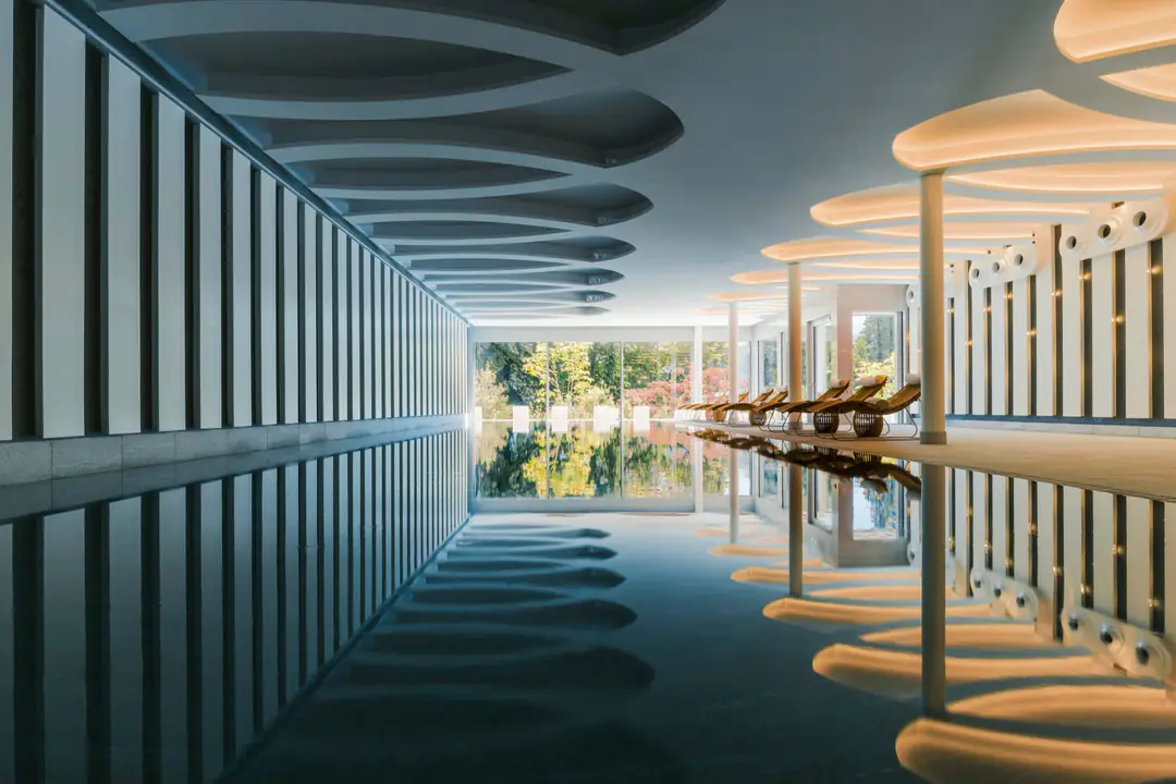 Long indoor pool with still dark water, lit wave-shaped ceiling and loungers beside garden windows