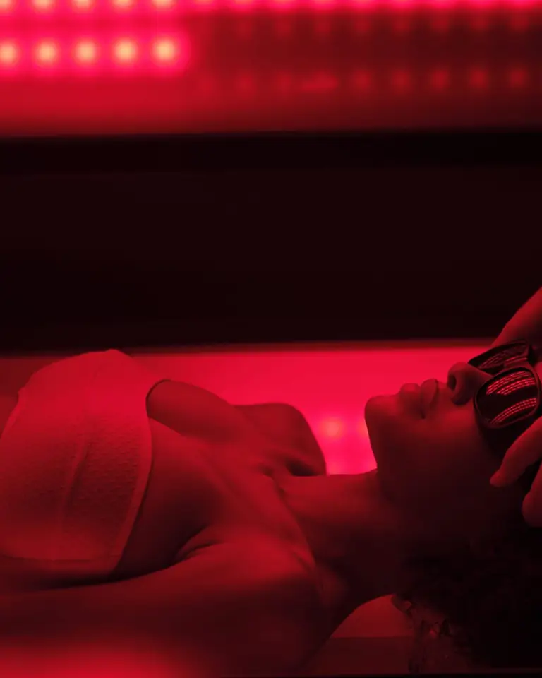 Guest in eye goggles lying in a whole-body photobiomodulation bed glowing red, a therapist's hands at her head