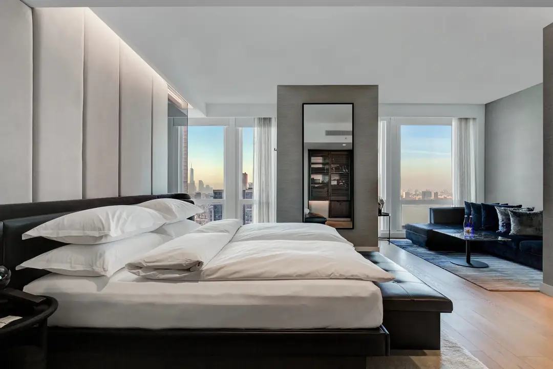 Premier King room with a low white bed, a mirrored partition and floor-to-ceiling windows over the Manhattan skyline