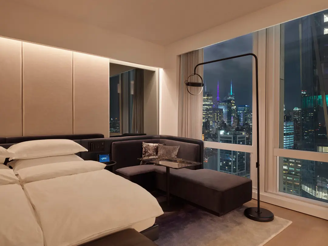 Dimly lit guest room at night with a white bed, a velvet banquette and a window onto lit Midtown towers