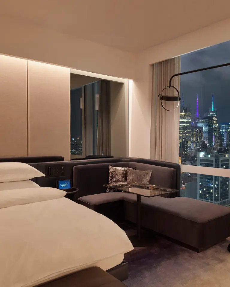 Dimly lit guest room at night with a white bed, a velvet banquette and a window onto lit Midtown towers