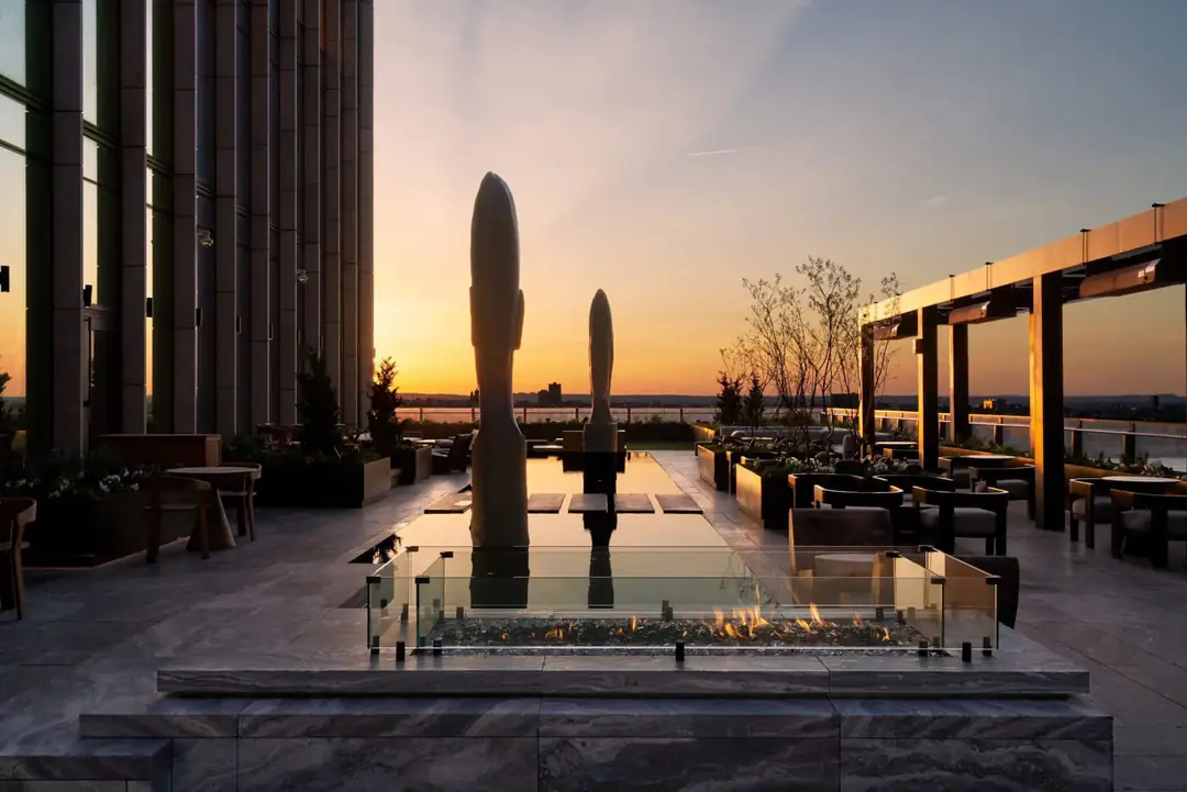 Rooftop terrace at sunset with a reflecting pool, two tall sculptures, a glass-walled fire pit and lounge seating