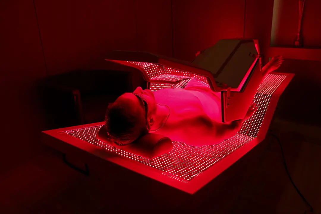 A man in goggles lies on a full-body LED bed glowing red, its hinged top panel raised