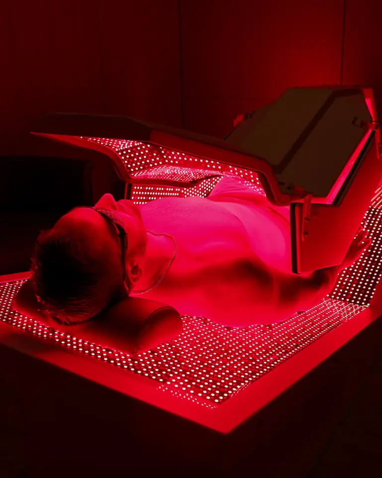 A man in goggles lies on a full-body LED bed glowing red, its hinged top panel raised