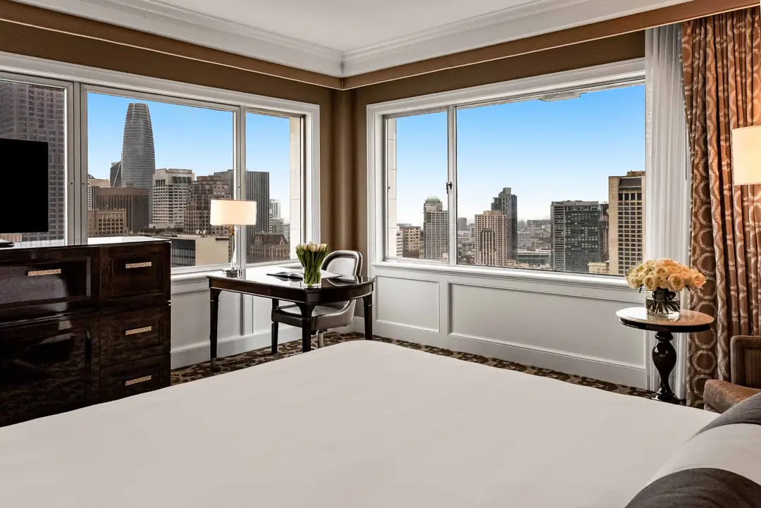 A corner guest room with two walls of windows onto downtown towers, a desk, dark dresser and white bed