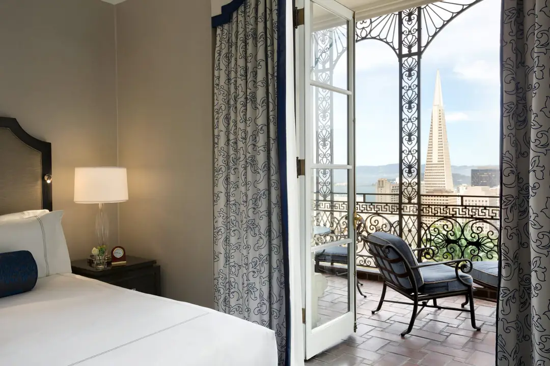 A bedroom with open French doors to a wrought-iron balcony looking at the Transamerica Pyramid and the Bay