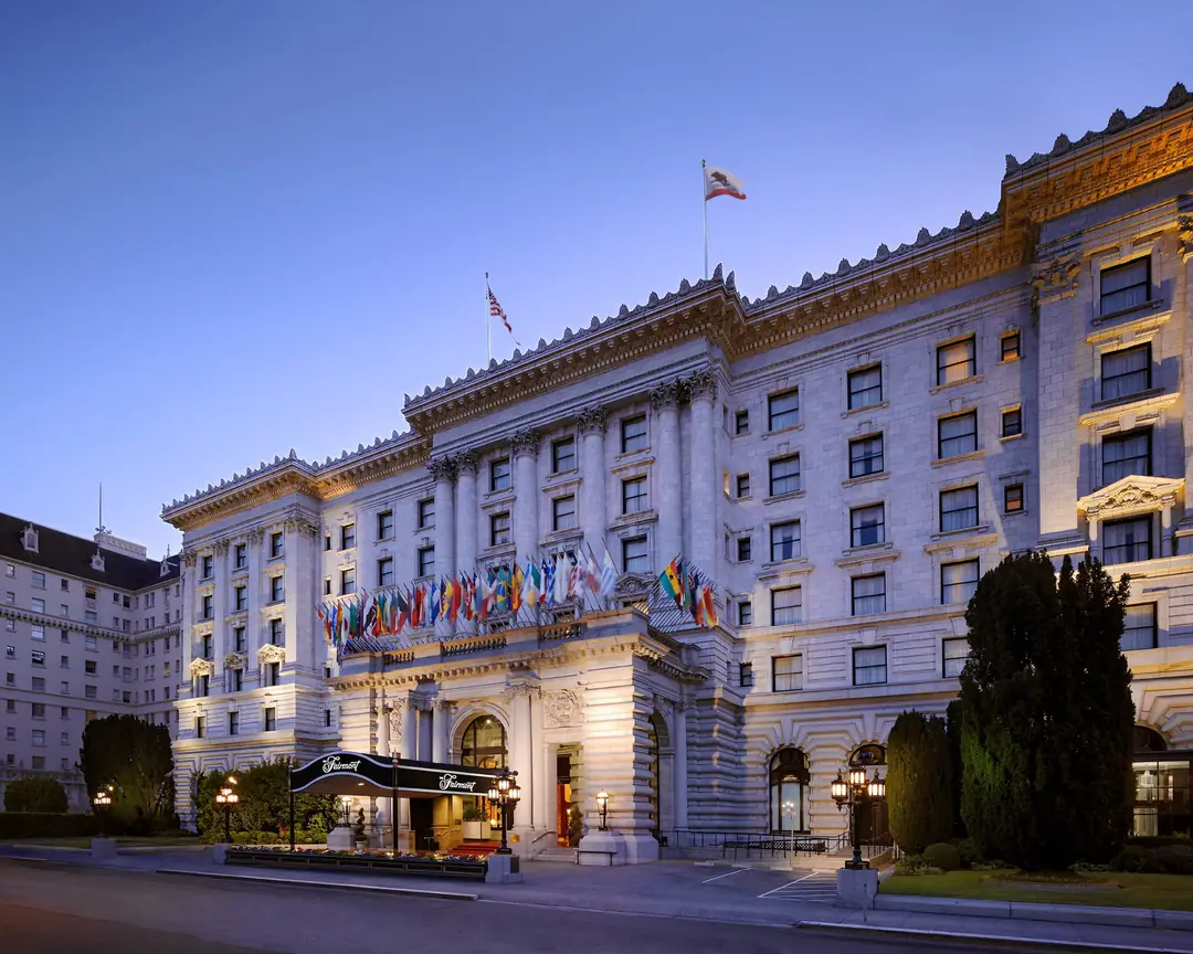 The Fairmont's white Beaux-Arts facade at dusk, with a row of flags over the lit entrance and black canopy