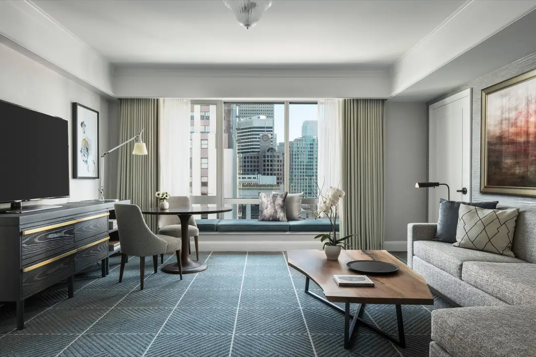 Suite living room in grey and blue, with a window seat, walnut coffee table and downtown towers outside