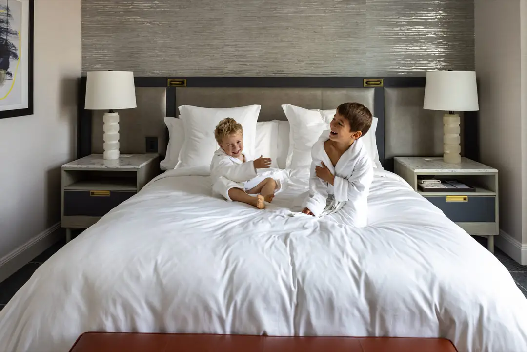Two children in white robes on a bed with white linen, between alabaster lamps and marble nightstands