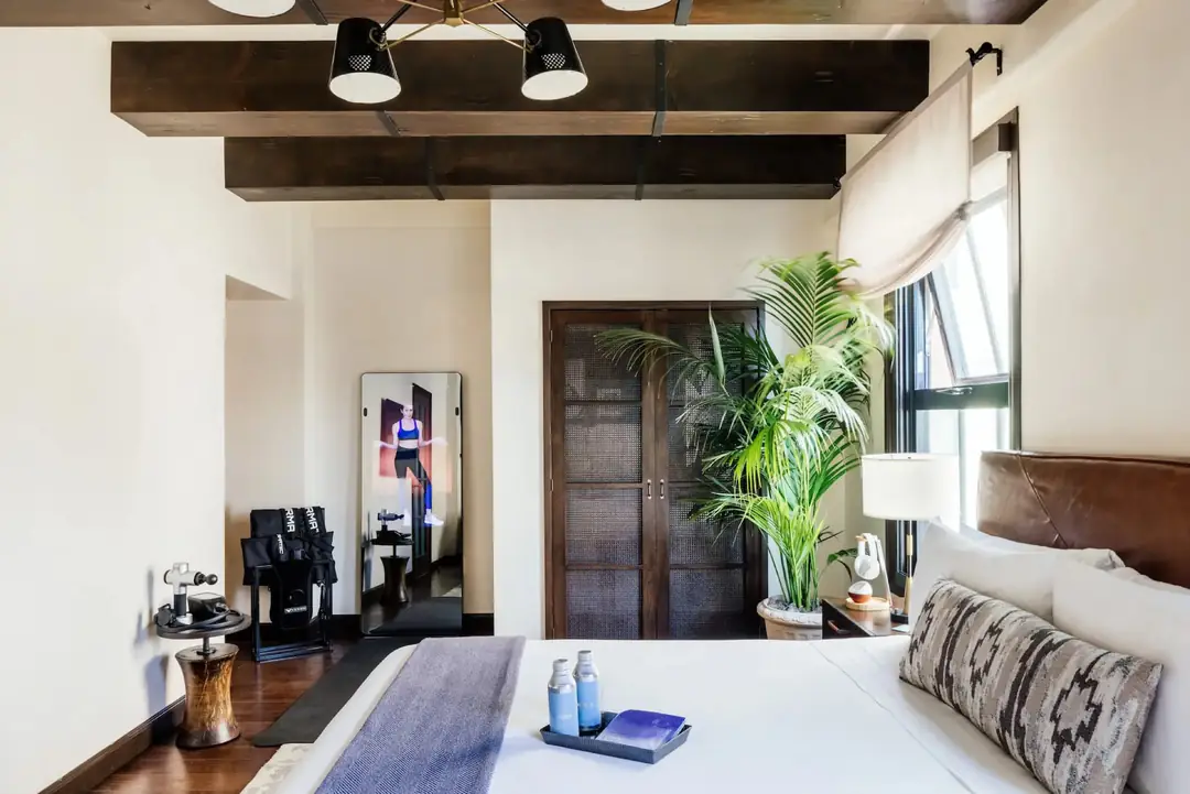 Rest & Recovery Suite bedroom: white bed with leather headboard, palm by the window, workout mirror on the far wall