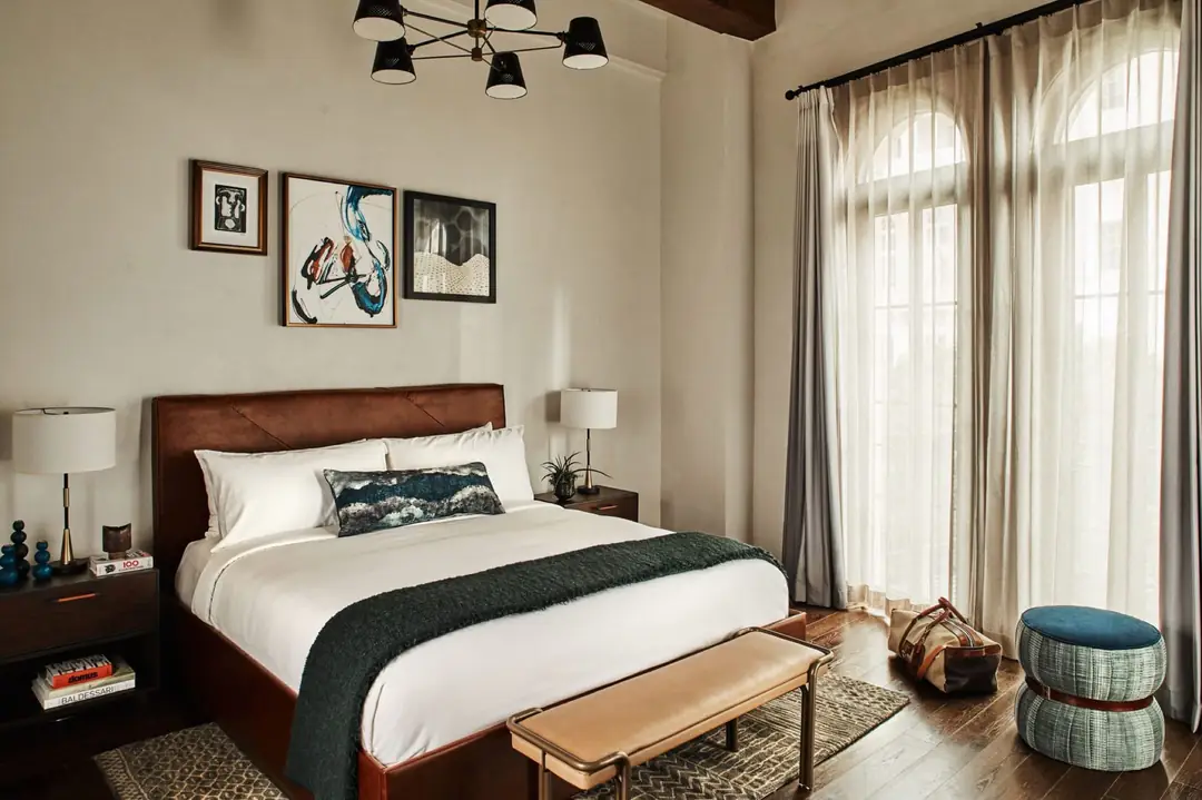 Suite bedroom with leather bed, green throw, wood floor and tall arched windows behind sheer curtains