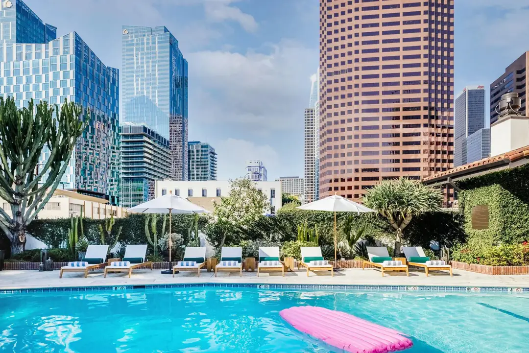 Outdoor pool with a pink float, white loungers, umbrellas and tall cacti below downtown Los Angeles towers