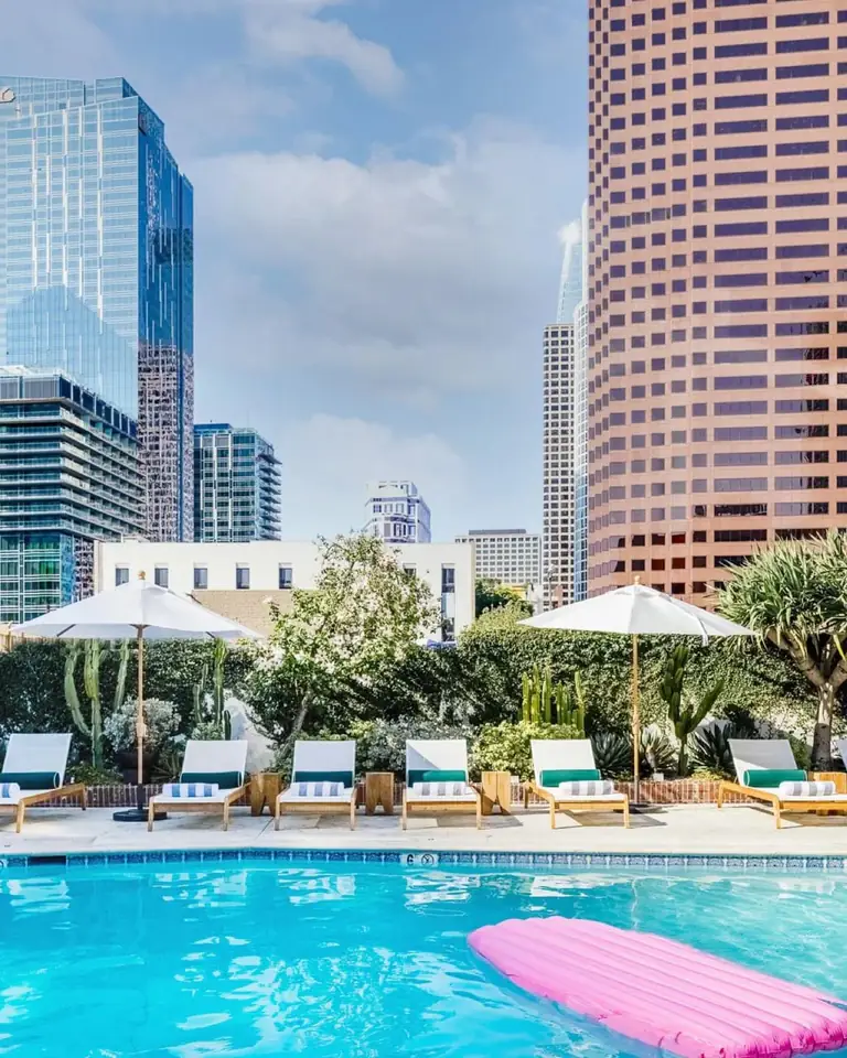 Outdoor pool with a pink float, white loungers, umbrellas and tall cacti below downtown Los Angeles towers