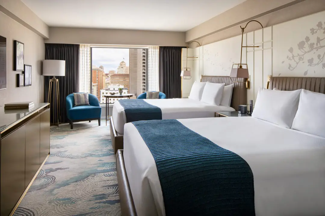 Guest room with two queen beds, blue throws, a cherry-blossom wall panel and a window onto city buildings