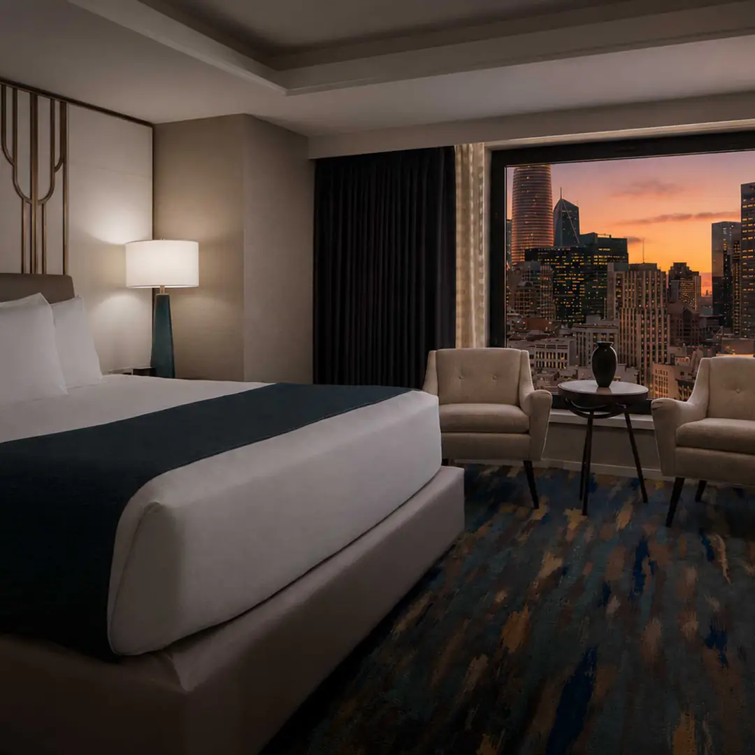 King bed and two armchairs facing a large window over the downtown skyline at dusk