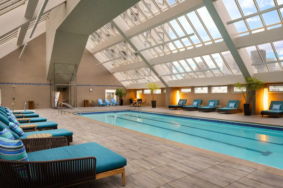 Indoor lap pool under a sloped glass atrium, teal loungers along both sides and city towers beyond
