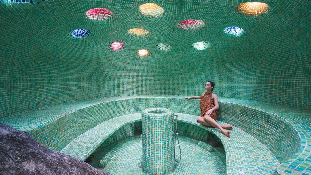 Green-tiled domed steam cavern with coloured glass skylights, a granite boulder and a guest on the bench