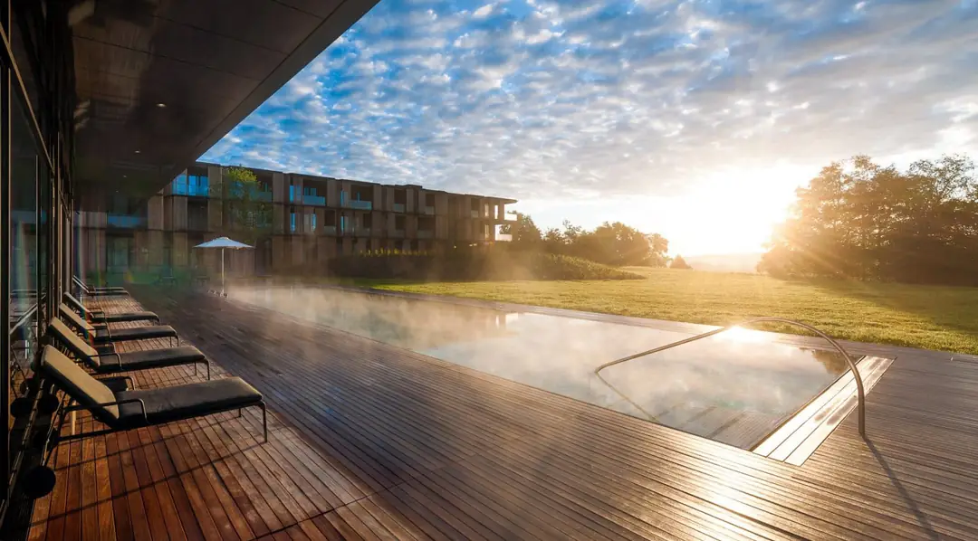 Steaming outdoor pool at sunrise, wooden deck with loungers, larch-clad wing and meadow beyond