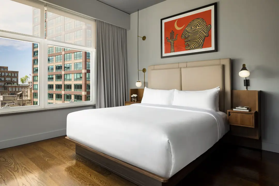 Guest room with a white bed, wood floor, wooden side tables, an orange painting and a large window