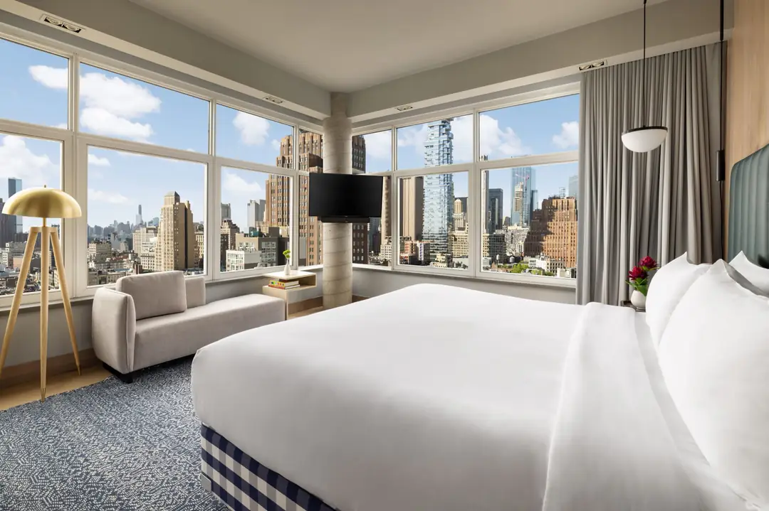 Corner bedroom of the Gallery Penthouse: a white king bed on a blue-check base, with floor-to-ceiling windows onto Manhattan