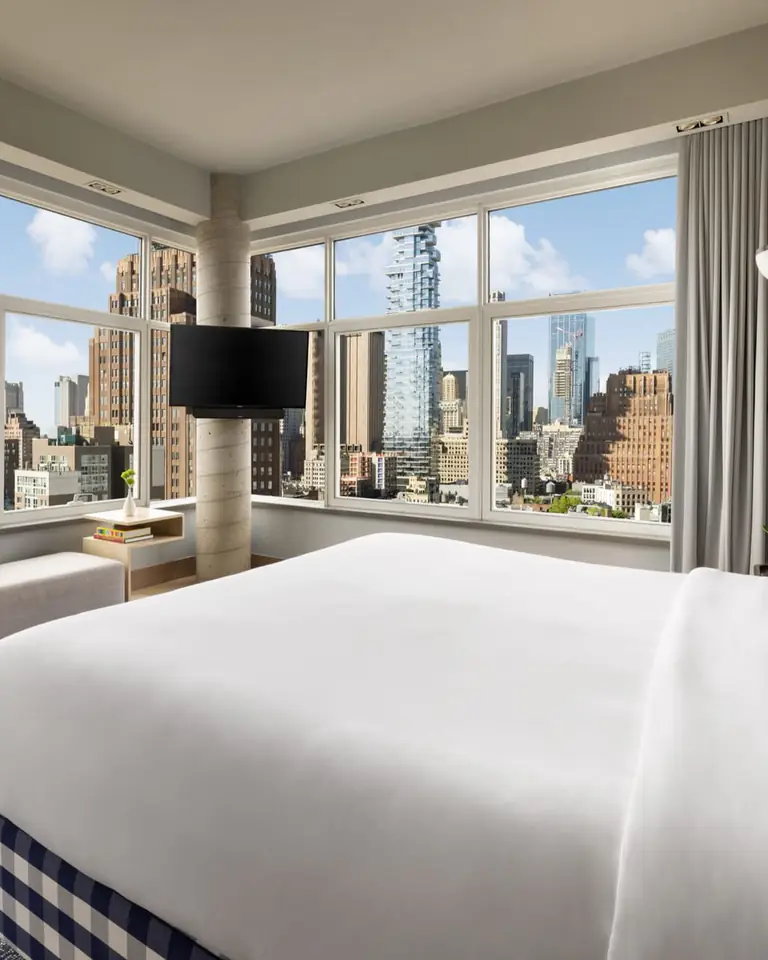 Corner bedroom of the Gallery Penthouse: a white king bed on a blue-check base, with floor-to-ceiling windows onto Manhattan