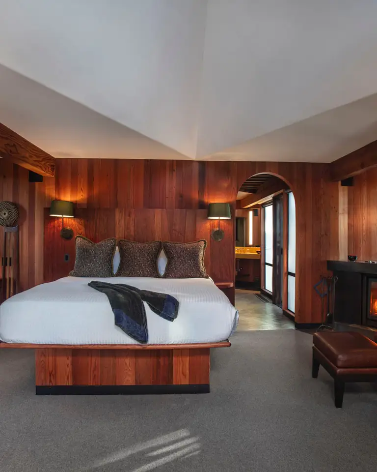 Tree House bedroom with redwood walls, a white bed, wood-burning fireplace and windows onto trees; no TV