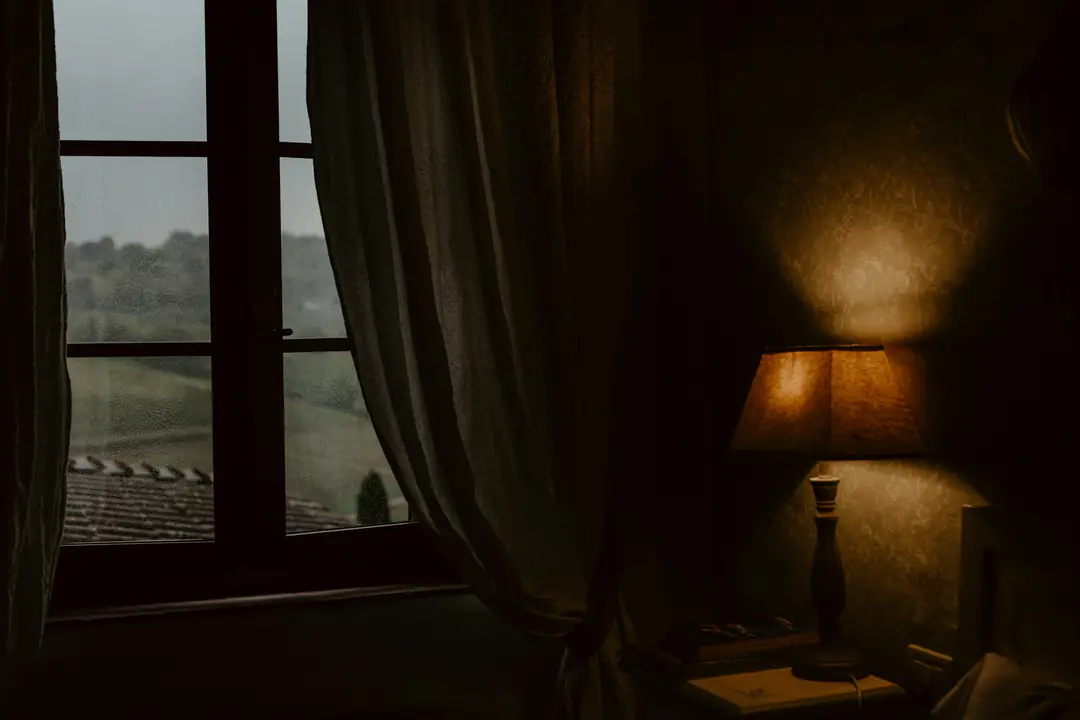 A lamp glowing beside a curtained window at dusk