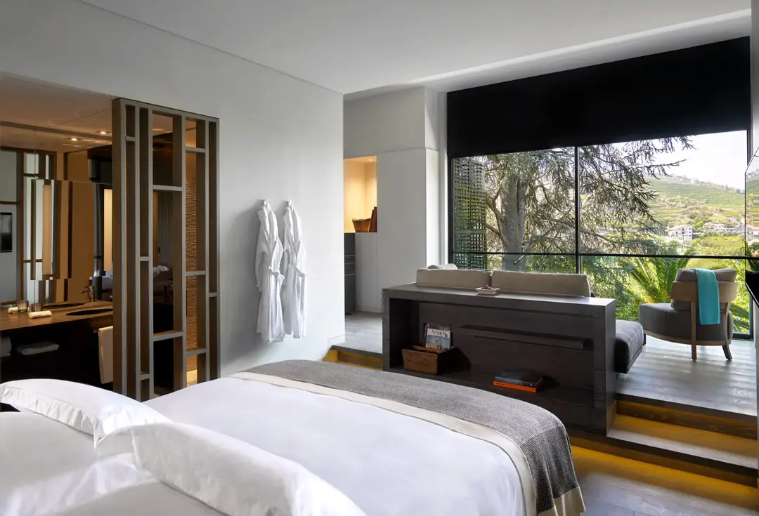 Quinta Deluxe room: white bed, wood floor, and a window seat facing a cedar and the valley