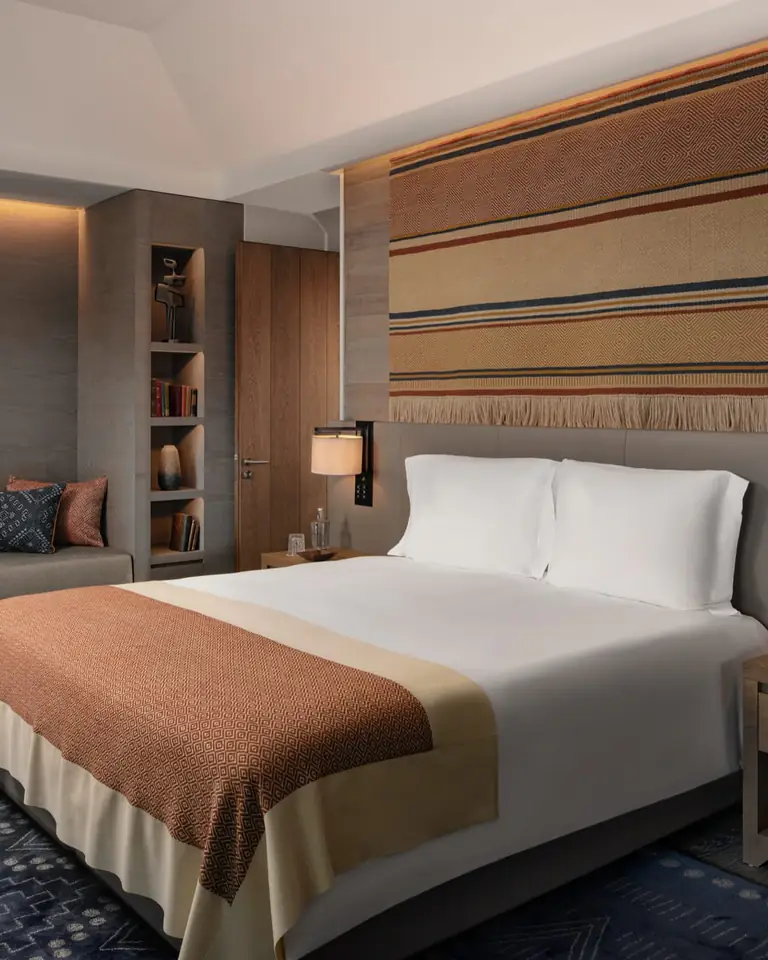 Bed with white pillows and a rust throw under a woven wall hanging, lit by two bedside lamps