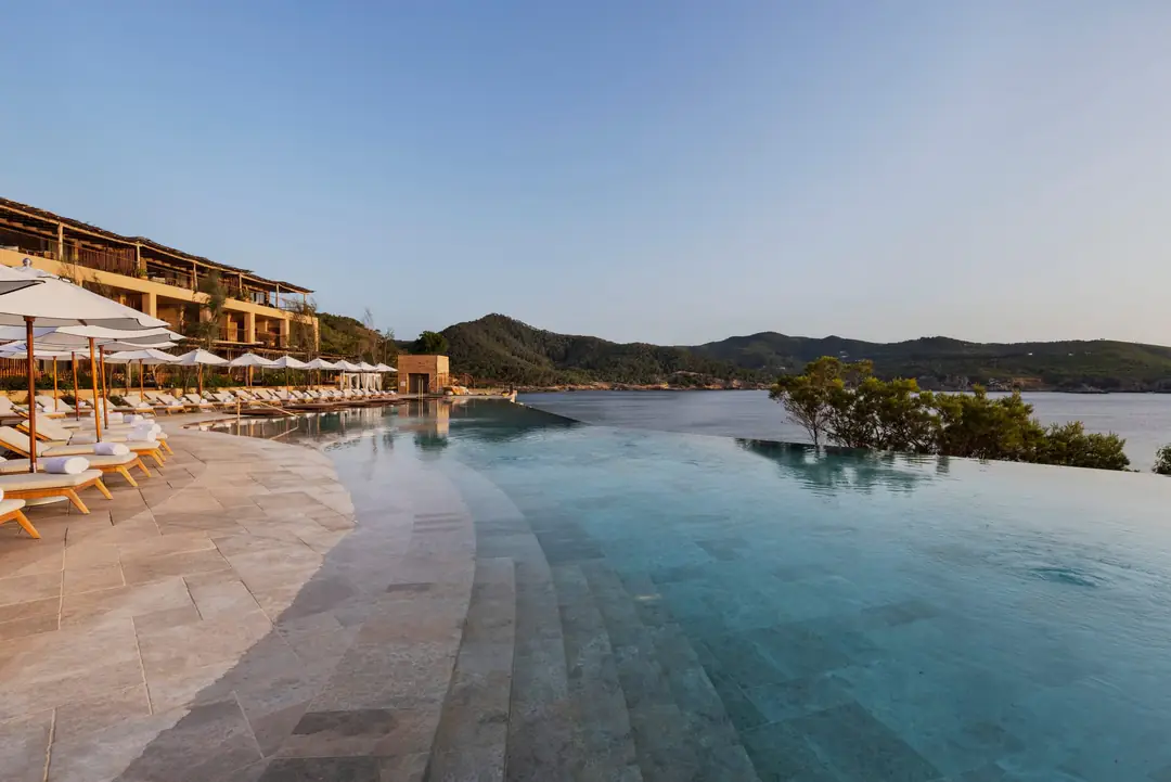 Infinity pool with stone steps and sun loungers, terraced resort buildings and the bay behind