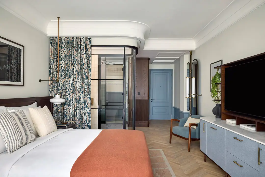 Bed in white linen with a rust throw, herringbone floor, blue cabinets and a glass-walled shower behind