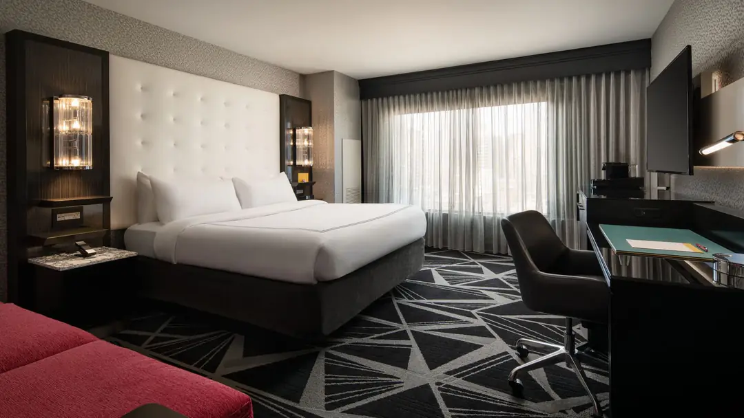 King guest room with a white tufted headboard, sheer-curtained window, desk and patterned carpet
