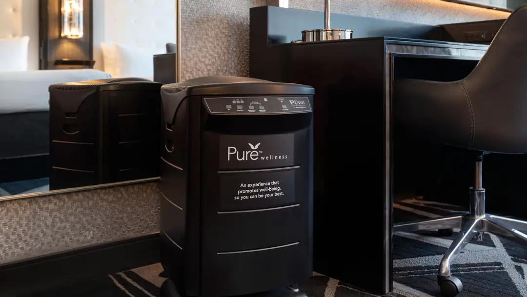 Black Pure Wellness air purifier on the floor beside a guest room desk and chair