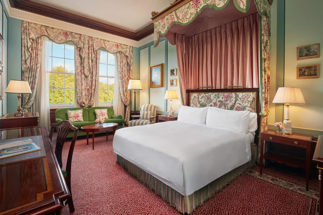 Guest room with a canopied double bed, floral curtains, red carpet and sash windows onto green trees